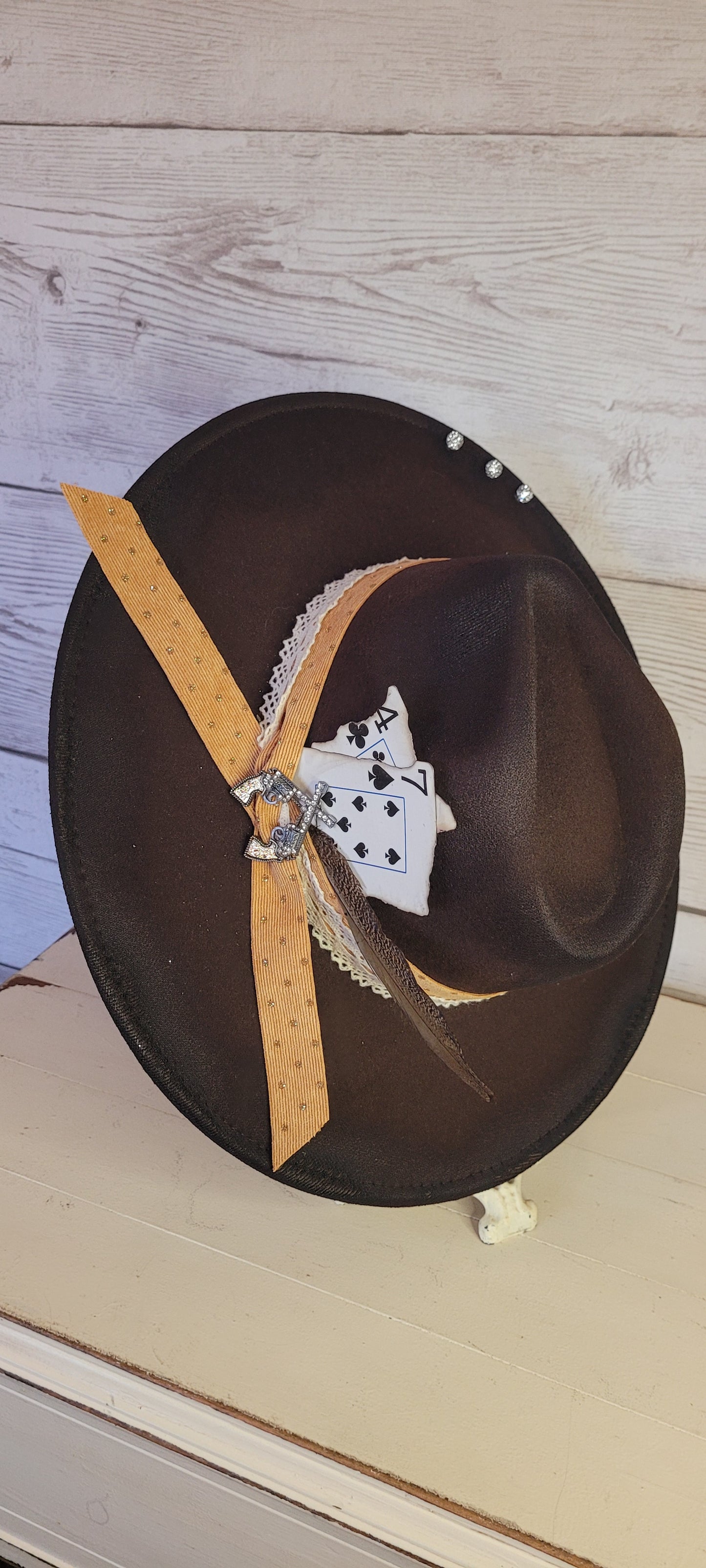Features natural lace ribbon, corduroy ribbon, feather, playing cards, pistol brooch, & rhinestone beads Felt hat Flat brim 65% polyester and 35% cotton Ribbon drawstring for hat size adjustment Head Circumference: 24" Crown Height: 5" Brim Length: 15.75" Brim Width: 14.5" Branded & numbered inside crown Custom designed by Kayla