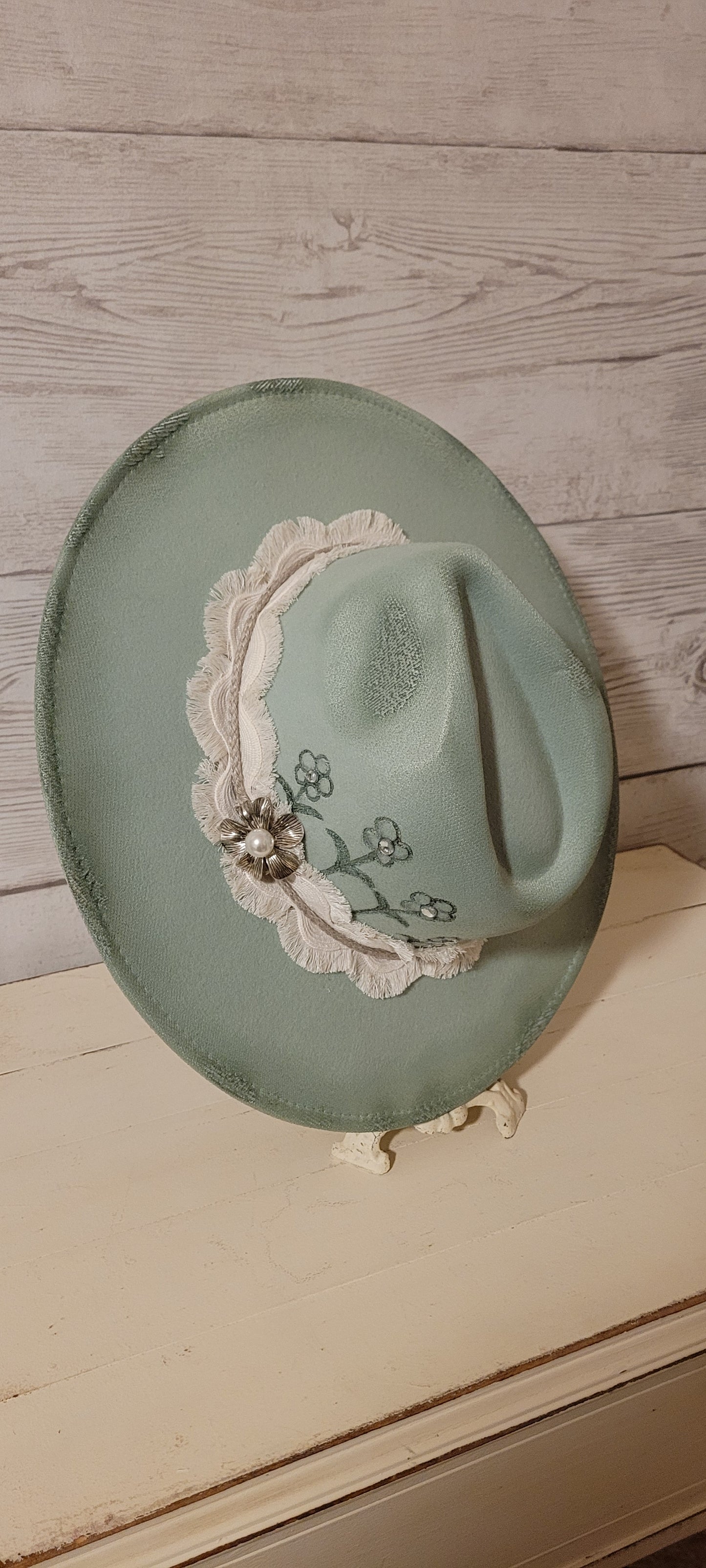 Features flowers engraved Wavy frayed ribbon, natural cord, rhinestones & metal pearl brooch Felt hat Flat brim 100% polyester Ribbon drawstring for hat size adjustment Head Circumference: 24" Crown Height: 5" Brim Length: 15.75" Brim Width: 14.5" Branded & numbered inside crown Custom burned & engraved by Kayla